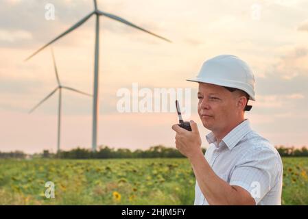 Bauarbeiter mit Walkie-Talkie gegen Windturbinenpark. Ein Mann mit weißem Helm, der ein Walkie-Talkie hält, um das Windturbinensystem zu überprüfen. Stockfoto