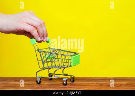 Weibliche Hand schieben Spielzeug Shopping-Trolley auf gelbem Hintergrund. Leerer Einkaufswagen mit Händen der Frau. Stockfoto