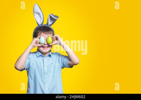 Niedlicher Junge mit Hasenohren, der farbige Eier neben den Augen auf gelbem Hintergrund hält. Fröhlicher Junge mit ostereiern und Hasenohren auf gelbem Hintergrund. Stockfoto