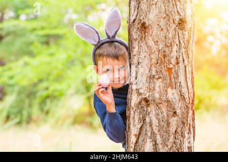 Ein süßer kleiner Junge mit einem Hasen-Ohr, der hinter einem Baum steht und das Osterei in der Hand hält. Frohe Ostern. Stockfoto