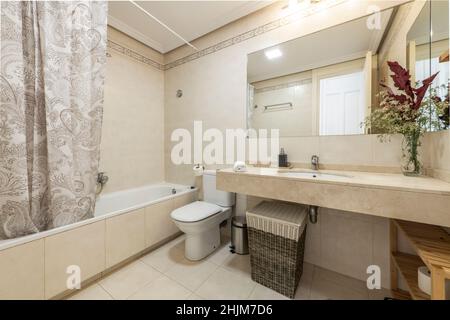 Badezimmer mit cremefarbener Marmorplatte, langer Badewanne mit Vorhang, Blumenstrauß, Porzellanwaschbecken und eingebautem Spiegel in der Apar der Ferienwohnung Stockfoto