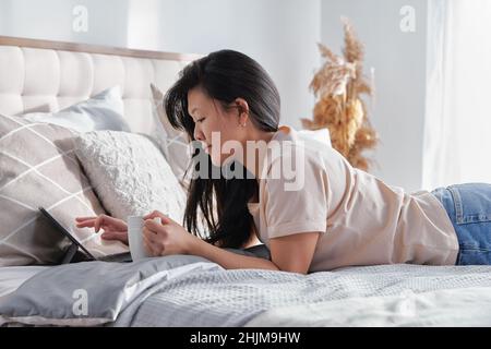 Seriöse Frau in legerer Kleidung hält Tasse arbeiten auf digitalen Tablet liegend auf dem Bett Stockfoto