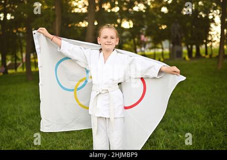DONEZK, UKRAINE - 10. JULI 2021. Ein Mädchen in einem weißen Kimono hält eine Flagge mit olympischen Symbolen in den Händen und lächelt. Stockfoto