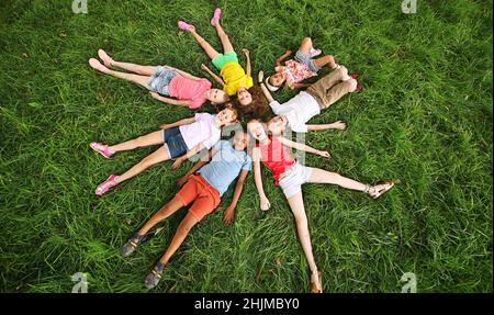 Kinder verschiedener Rassen und Nationalitäten liegen in farbenfrohen Kleidern auf dem Gras und lächeln. Stockfoto