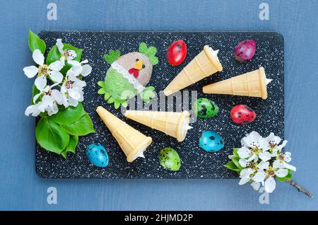 Ostern buntes Stillleben auf einem Schneidebrett mit bunten Wachteleiern, Dessert in Waffelkegel und einem Spielzeug-Huhn auf einem Hintergrund von Fruchtinflorescen Stockfoto