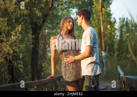 Ein schönes Paar in der Natur steht umarmt auf einer Holzbrücke über einen mit Schilf bewachsenen Fluss. Glückliche Liebhaber feiern den Valentinstag gemeinsam. Stockfoto