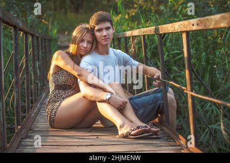Ein Mann und ein Mädchen sitzen auf einer Holzbrücke über einen Fluss in einem Stadtpark. Liebhaber sitzen umarmt in der Natur Stockfoto