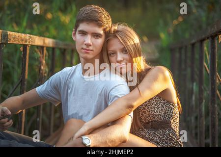 Ein hübscher Kerl sitzt und umarmt ein minderjähriges Mädchen auf einer Brücke mit rostigen Geländern. Ein glückliches junges Paar in der Natur kommuniziert und verbringt Zeit miteinander. Stockfoto