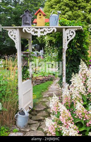 Steinweg durch weiße Holzarbour mit Rollwerk, Vogelhaus und Weinlese Gießkanne von Hydrangea 'Pinky Winky' begrenzt dekoriert. Stockfoto