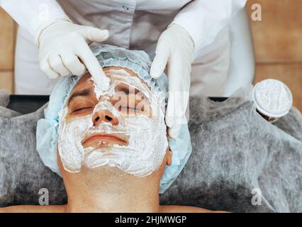 Kosmetikerin Anwendung Peeling-Maske auf das Gesicht zu jungen Mann, Spa Schönheitssalon. Das Konzept der Körper- und Gesichtspflege für Männer Stockfoto