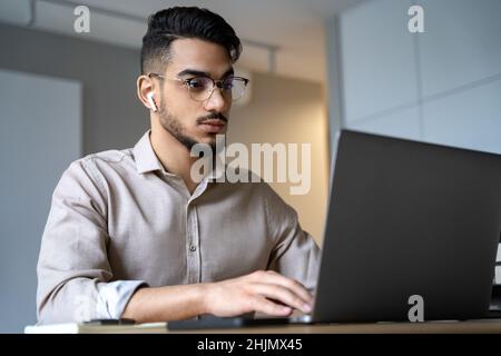 Porträt eines jungen indischen Mannes, der auf den Laptop-Bildschirm schaut, der im Heimbüro sitzt Stockfoto