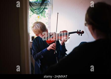 Kleiner Junge, der seine Geige spielt, während seine stolze Mutter zuschaut. Stockfoto