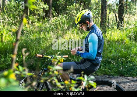 Ein männlicher Mountainbiker mit seinen 30s und 40s, der mitten im Wald auf seinem Fahrrad eine App auf seinem Smartphone überprüft Stockfoto