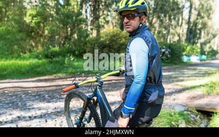 Porträt eines Mountainbiker, der mit Helm, Brille und Smartwatch auf seinem Fahrrad auf einer von Bäumen gesäumten Landstraße mit Kopierfläche sitzt Stockfoto