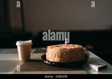 Kuchen mit einer Kerze und einer Papiertasse mit Kaffee in einem Café. Frühstück Americano in einem Glas und einem festlichen Dessert. Geburtstagskuchen napoleon. Café-Menü Stockfoto