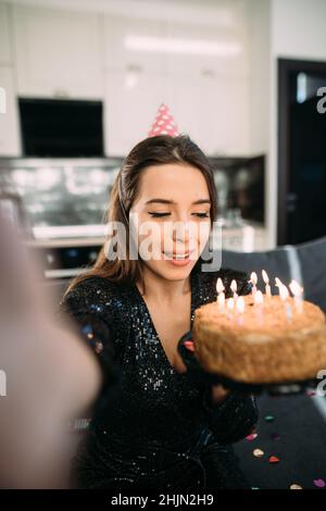 Lächelnd positive junge Frau feiert Geburtstag zu Hause, hält Kuchen mit Kerzen, Mädchen 25 Jahre alt feiert einen Feiertag, alles gute zum Geburtstag Stockfoto