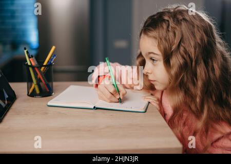 Mädchen sitzt am Schreibtisch und schaut sich den Online-Kurs auf dem Tablet an, während sie in ihr Notebook schreibt. Online-Lernschule. Online-Unterricht. Stockfoto