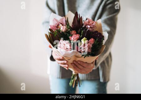 Junge Frau in grauem Strickpullover mit Bouqet aus verschiedenen rosa Blumen in den Händen auf dem grauen Hintergrund, selecrive Fokus Stockfoto
