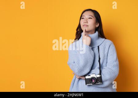 Junge asiatische Frau mit einer Kamera um den Hals und nachdenklichem Gesichtsausdruck mit orangefarbenem Hintergrund Stockfoto