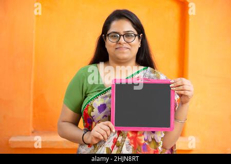 Indische Frau trägt Saree und Brille mit Schiefer vor orangefarbenem Hintergrund, lächelnde asiatische Lehrerin mit schwarzem Kreidetafel. Bildung konz Stockfoto