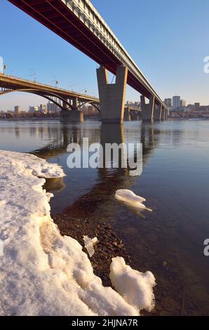 Metro-most und Automobilbrücke über den großen sibirischen Fluss in Nowosibirsk. Schnee und Eis am Strand im frühen Frühjahr. Hohe Gebäude auf urbanem Boden Stockfoto