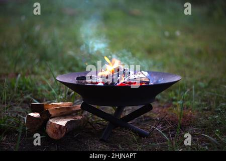 Die Flamme des Feuers brennt in einer Metall-Feuerschale - erwärmen Sie Ihre Hände am Feuer, rühren Sie das Feuerholz mit einem Stock. Eine Feuerstelle mit Feuerholz für sicheren Campfi Stockfoto