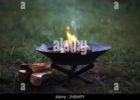 Die Flamme des Feuers brennt in einer Metall-Feuerschale - erwärmen Sie Ihre Hände am Feuer, rühren Sie das Feuerholz mit einem Stock. Eine Feuerstelle mit Feuerholz für sicheren Campfi Stockfoto