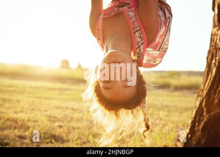 Das Mädchen hängt im Sommer kopfüber auf einem Baum in orangefarbenem Sonnenlicht und einem leichten Kleid. Sommerzeit, Hitze, Kindheit. Lustige Porträt mit zerzaust Stockfoto