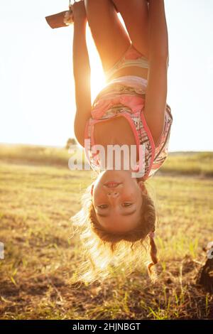 Das Mädchen hängt im Sommer kopfüber auf einem Baum in orangefarbenem Sonnenlicht und einem leichten Kleid. Sommerzeit, Hitze, Kindheit. Lustige Porträt mit zerzaust Stockfoto