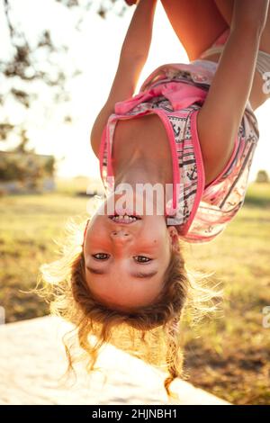 Das Mädchen hängt im Sommer kopfüber auf einem Baum in orangefarbenem Sonnenlicht und einem leichten Kleid. Sommerzeit, Hitze, Kindheit. Lustige Porträt mit zerzaust Stockfoto