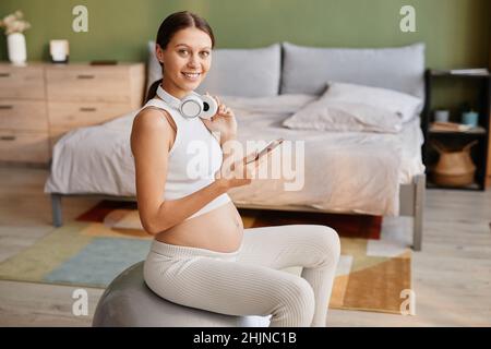 Porträt einer jungen Schwangeren in Kopfhörern, die die Kamera lächeln, während sie zu Hause Musik hören und auf dem Fitnessball trainieren Stockfoto