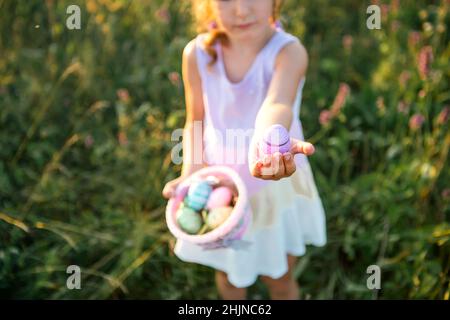Nettes lustiges Mädchen mit bemalten Ostereiern im Frühling in der Natur auf einem Feld mit goldenem Sonnenlicht und Blumen. Osterferien, Osterhase mit Ohren, Colo Stockfoto