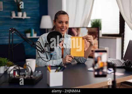 Influencerin präsentiert gelbes Buch vor dem Aufnahmemoster des Smartphones. Similing Content Creator sendet Review für Social Media-Kanal im Heimstudio mit Mikrofon und Laptop. Stockfoto