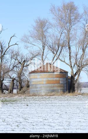 Getreideeimer mit Schnee und blauem Himmel und Bäumen auf einem Farmfeld Stockfoto