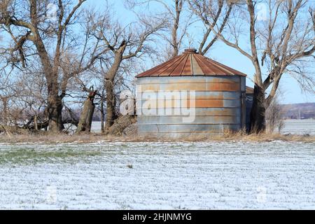 Getreideeimer mit Schnee und blauem Himmel und Bäumen auf einem Farmfeld Stockfoto