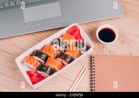 Verschiedene Sushi-Rollen mit Lachs in Lieferkarton auf Holztischhintergrund mit Laptop. Online-Home-Bestellung von Lebensmitteln und Lieferung Food-Konzept Stockfoto