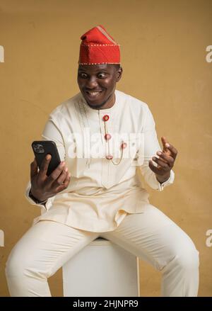 Yoruba kulturell gekleideter Geschäftsmann, der mit dem Telefon in der Hand sitzt, ist aufgeregt Stockfoto