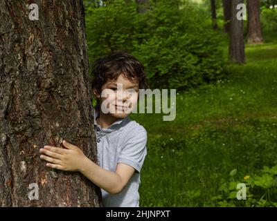 Lächelnder Junge umarmt Baum in einem Wald. . Nahaufnahme eines sorglosen Teenagers. Stockfoto
