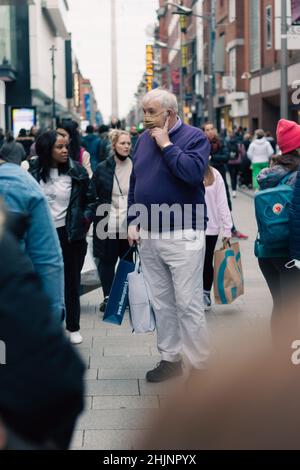 Große alte Männer mit Gesichtsmaske zwischen Menschenmengen an der Henry Street mit Einkaufstasche in der Hand, verschwommene Straßenfotografie im Hintergrund, Dublin, Irland Stockfoto