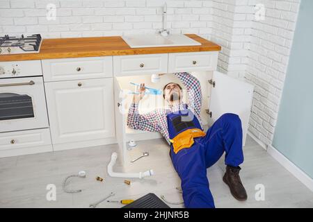Männlicher Klempner in Arbeitskleidung repariert und unterhält Siphon unter dem Waschbecken in der Küche der Wohnung. Stockfoto