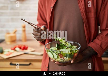 Nahaufnahme eines jungen schwarzen Mannes, der Salat in einer Glasschüssel mischt, während er in einer gemütlichen Küche gesunde Mahlzeiten genießt, Platz zum Kopieren Stockfoto
