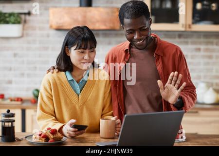 Waist-up-Porträt eines glücklichen Mixed-Race-Paares, das im Videochat in der gemütlichen, farbenfrohen Küche mit der Kamera winkt Stockfoto