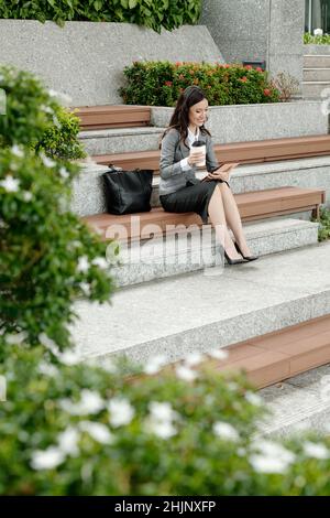 Unternehmerin sitzt auf der Treppe, trinkt Kaffee und liest Nachrichtenartikel auf Tablet-Computer Stockfoto