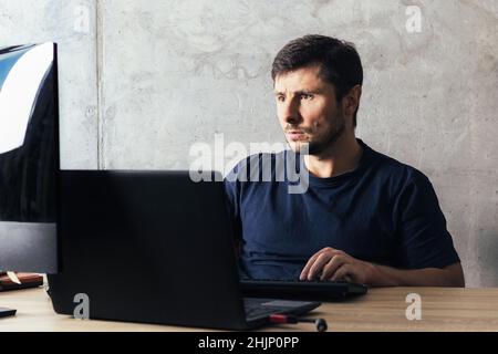 Ein hübscher, ernsthafter bärtiger Mann, der am Schreibtisch sitzt, an einem Laptop arbeitet und im modernen Büro den Computerbildschirm gegen eine Betonwand blickt Stockfoto