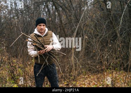 Porträt eines gefrorenen Touristen-Mannes, der warme Kleidung trägt und trockenes Totholz für das Feuer im Wald an einem bewölkten, kalten Tag sammelt. Stockfoto