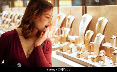 Überrascht hübsche Frau, die Schmuck im Schaufenster ansah. Mädchen in der Nähe von Schmuck. Interessierte Kunden wählen Gold, Diamanten oder Edelsteine. Einkäufern Stockfoto