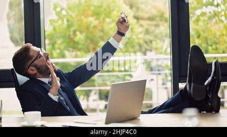 Aufgeregter Geschäftsmann, der mit Beinen auf dem Schreibtisch sitzt und mit der Hand winkt, während er Selfie auf dem Smartphone nimmt Stockfoto
