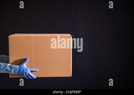Nahaufnahme der weiblichen Hände in Handschuhen und einem Jeanshemd. Der Lieferer übergibt den Karton auf schwarzem Hintergrund an den Kunden. Antimikrobiell Stockfoto