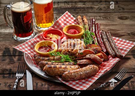Ein köstliches Set aus verschiedenen Bratwürsten, serviert mit Kartoffelkeilen im Landhausstil, gedünstetem Kohl und Saucen, die mit frischem Gemüse als Bier serviert werden Stockfoto