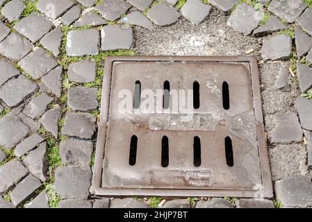 Altes Sturmgitter auf der Straße. Abwasserkanal von Roman Rain. Ein alter Steinpflasterstein hat ein eingebautes Metallgitter zum Sammeln von Regenwasser. Stockfoto